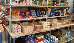 A stack of shelves is stocked with groceries and essentials like eggs, bread and rolls, water, juice, cooking accessories, paper towels and kitchen foil. On top of the stack of shelves are three folding camp chairs and table set up as a display.
