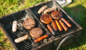 Sausages and burgers cook on a charcoal grill on the grass in the sunshine