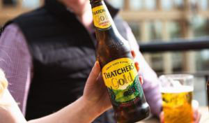 Someone holds a bottle of Thatcher’s Cider, which has a yellow label with the company branding on it. Someone behind is holding a Thatcher’s branded pint glass filled with golden cider.