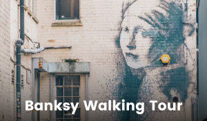 A woman stands in a courtyard in Bristol surrounded by white-painted brick buildings. There is a large Banksy graffiti work on the wall behind her depicting a version of the Girl With a Pearl Earring painting by Vermeer. Insead of a pearl earring, she wears an orange security alarm box