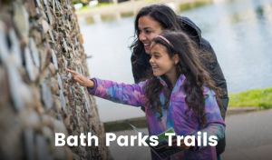A woman and girl are smiling as they study an ornamental stone wall for clues while on a treasure hunt in Bath. The girl is holding a pamphlet while pointing at something on the wall.