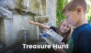 A girl and a boy stand in front of a historical fountain. The girl is smiling and pointing at something on the fountain feature, and the boy is looking at a pamphlet.