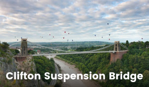 The iconic Clifton Suspension Bridge in Bristol spans the estuary. It’s two large pillars on either end support cables and metal work that suspend the bridge’s surface high above the water. There are hot air balloons scattered across the sky behind the bridge.