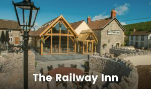 An old stone building stands to the right, with a modern oak-framed country-style extension to the left. There is a large patio out front with tables and chairs. The pub entrance porch has a smart thatched roof, and a sign hangs on the older building with the text ‘Railway Inn’ in black lettering on a cream background.