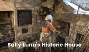 A mannequin in historic clothing stands in front of a bread oven, with a long-handled paddle used for removing baked goods from commercial ovens. The room is built of stone, with multiple ovens, fireplaces, a table and a variety of historical baking implements.
