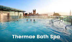 A rooftop pool filled with water from a thermal spring steams while four people swim and relax. The sky is clear and historic buildings of Bath are visible on the skyline